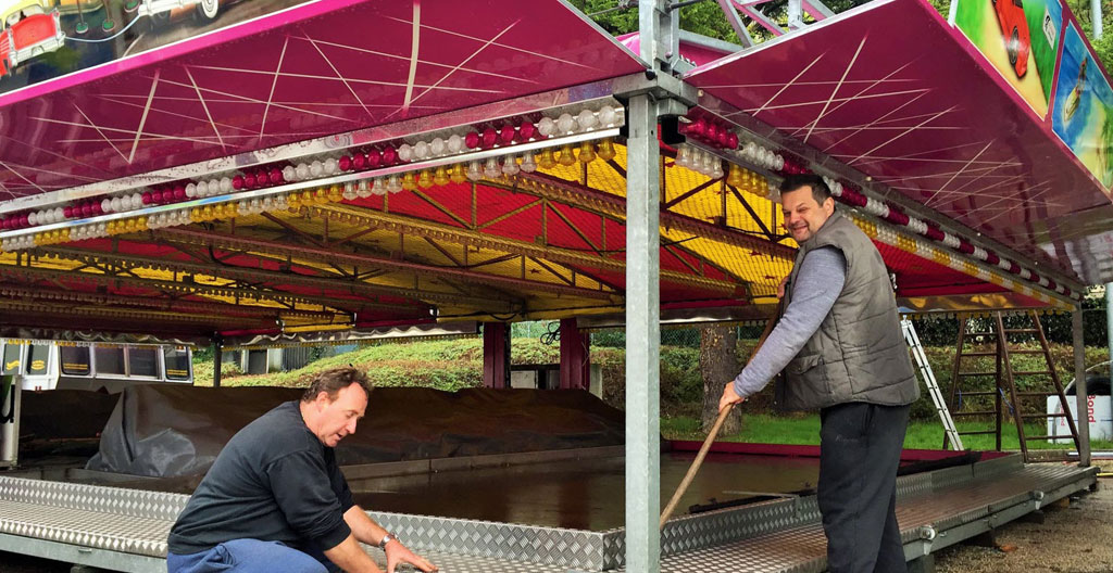 Techniciens installant et calant un carrousel ancien sur une esplanade événementielle, outils professionnels visibles, périmètre de sécurité matérialisé avec barrières, vue large montrant les contraintes d'espace